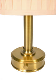 Close-up of a cordless table lamp with a round brushed brass base and a pleated light beige fabric shade by modern lantern