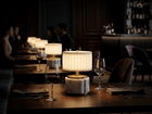 Nora rechargeable mini lamp with antique brass pleated shade on dining table in a dimly lit restaurant setting.