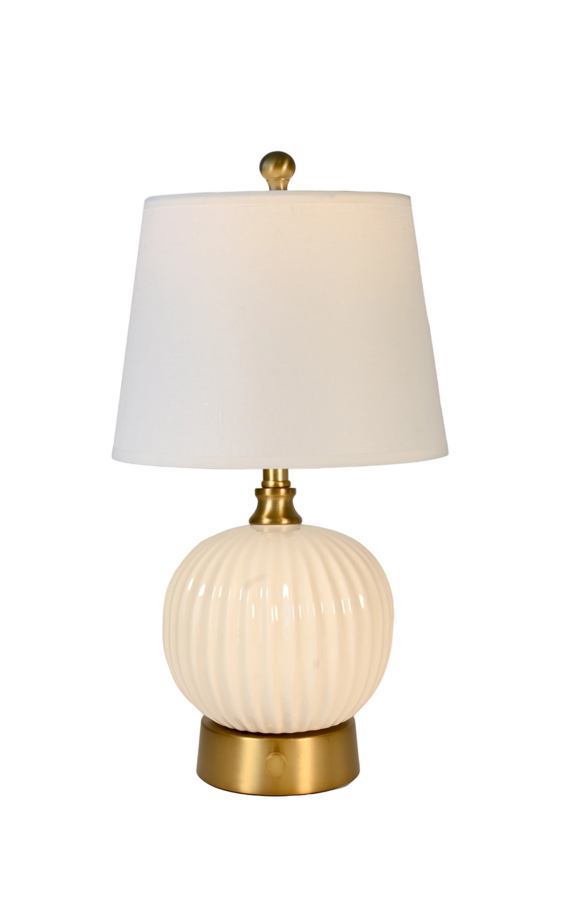A cordless table lamp with a ribbed cream-colored ceramic globe base, a white fabric drum shade, and antique brass gold-finished metal accents.
