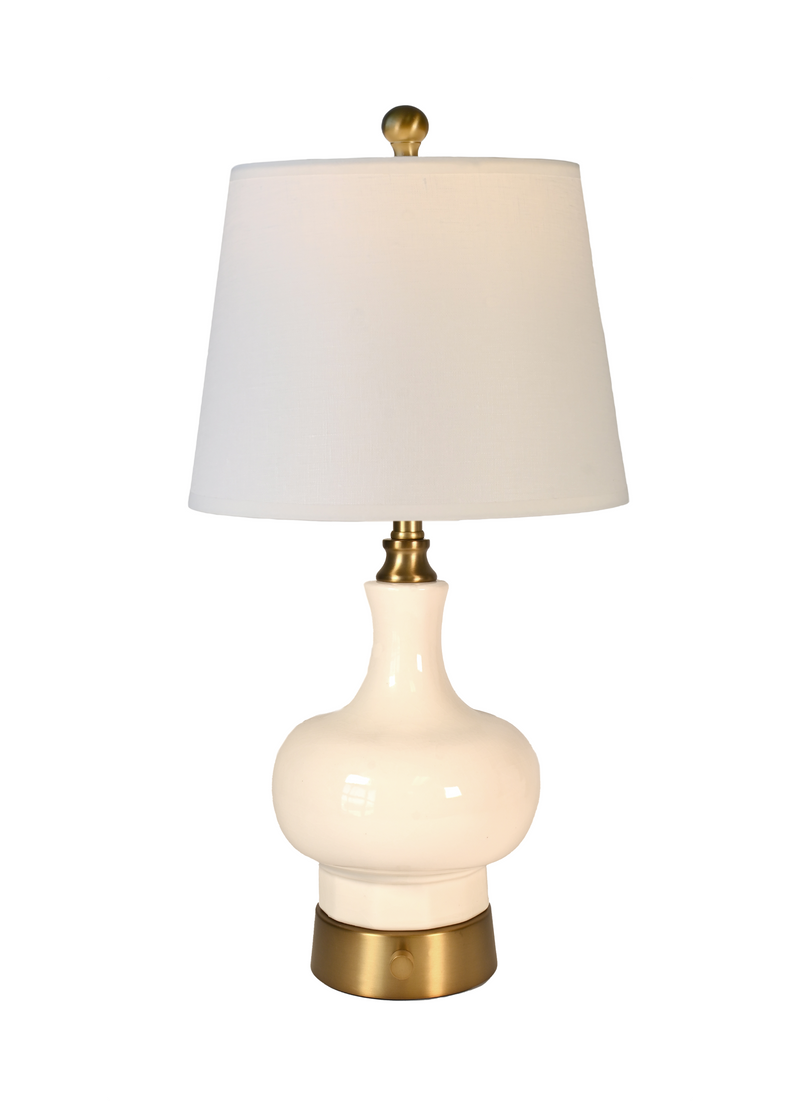 A cordless table lamp with a glossy cream ceramic bulb-shaped base, an off white fabric drum shade, and brass-finished metal accents at the top finial, neck, and bottom metal base.