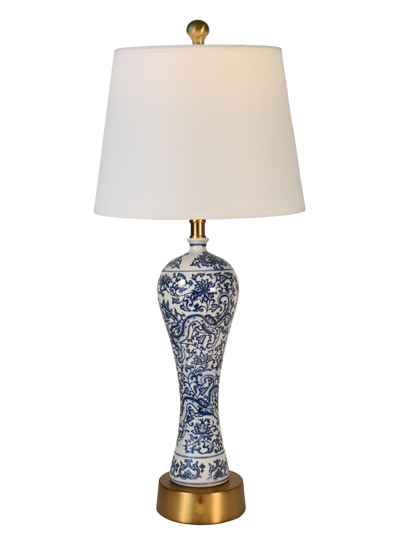 A hand painted blue and white table lamp with an off white fabric drum shade, a blue and white porcelain vase-shaped base, and an antique brass-finished metal top and base.
