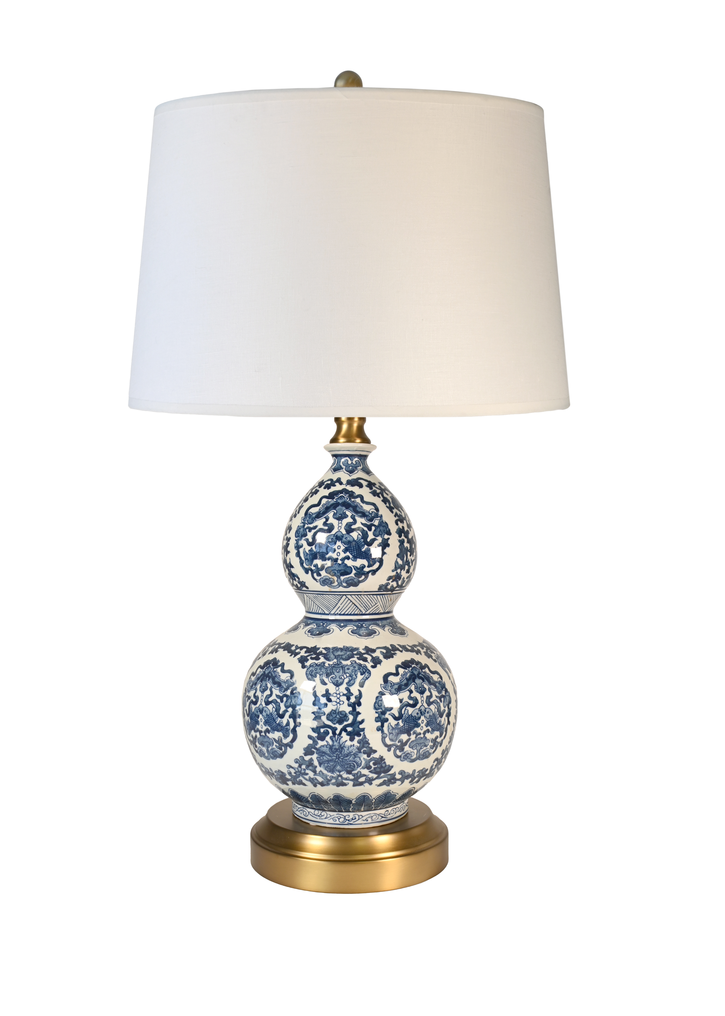 A cordless table lamp with a white drum shade, a blue and white porcelain gourd-shaped ceramic base, and a brass-finished metal neck and circular base.