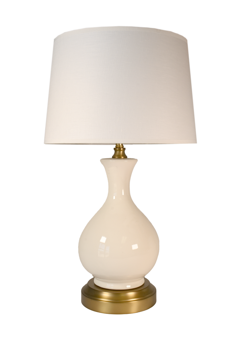 A cordless table lamp with a glossy cream-colored ceramic vase-shaped base, an off white linen shade, and a polished brass-finished metal top and circular base.