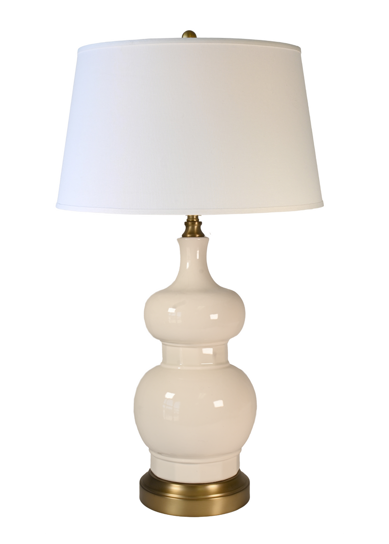 A table lamp with a glossy cream ceramic gourd-shaped base, an off white fabric drum shade, and  brass-finished metal accents at the top and bottom.
