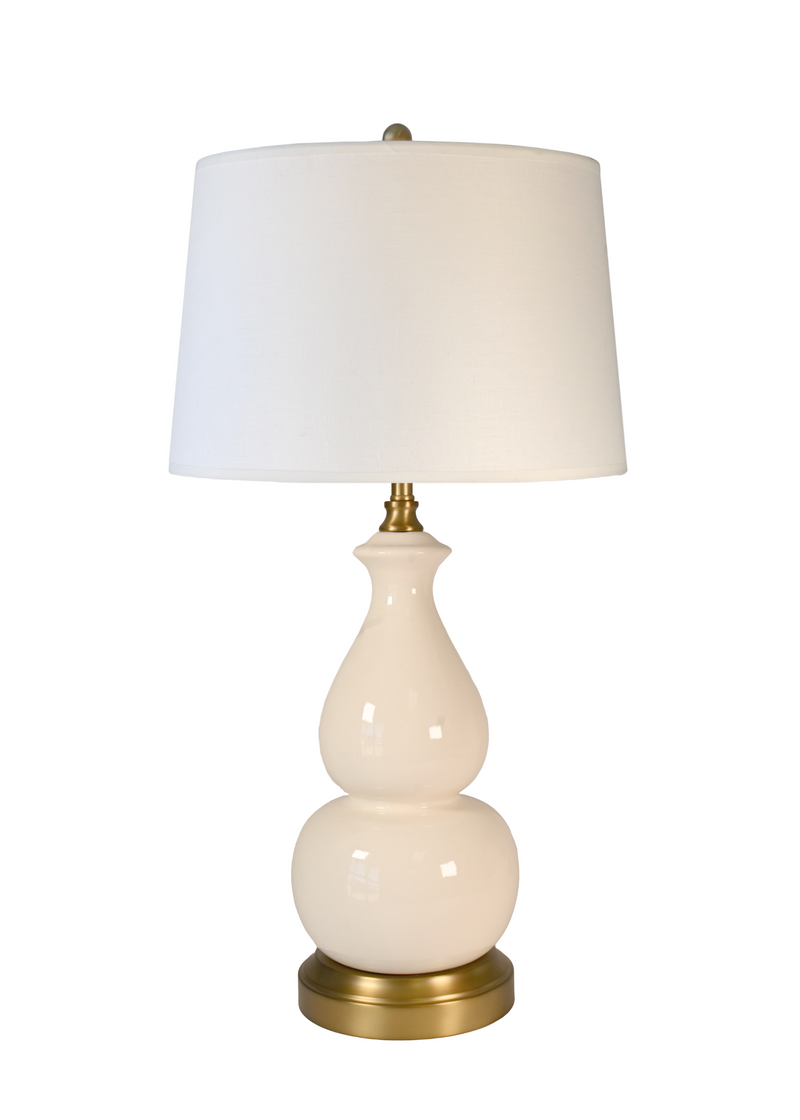 A cordless table lamp with an off white drum shade, a glossy cream gourd-shaped ceramic base, and a brass-finished neck and base, shown against a plain white background.