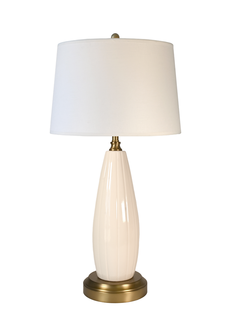 A cordless table lamp with an off white drum shade, a glossy cream ribbed ceramic base, and a brass-finished metal neck and circular base
