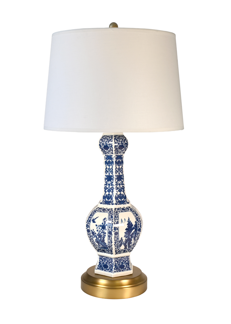 A cordless table lamp with an off white drum shade, a blue and white porcelain vase-shaped base, and a gold-finished metal base and finial, shown on a plain white background.