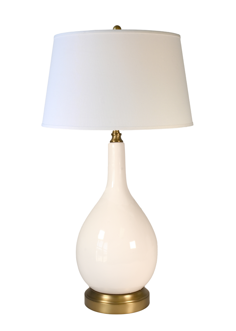 A large cordless table lamp with a glossy ivory teardrop-shaped ceramic base, an off white drum shade, and brass-finished metal accents at the neck and bottom.