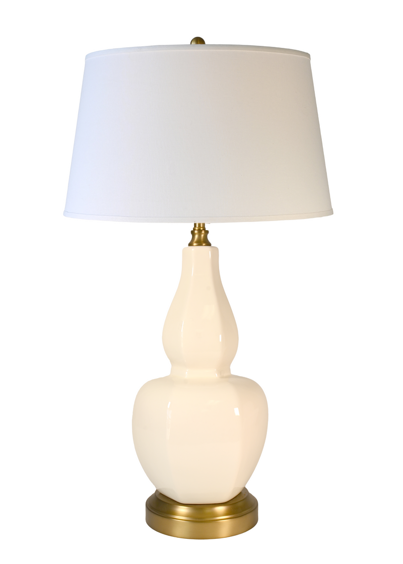 A cordless table lamp with a glossy cream-colored ceramic gourd-shaped base, an off white fabric drum shade, and a brass-finished metal accents at the neck and bottom.