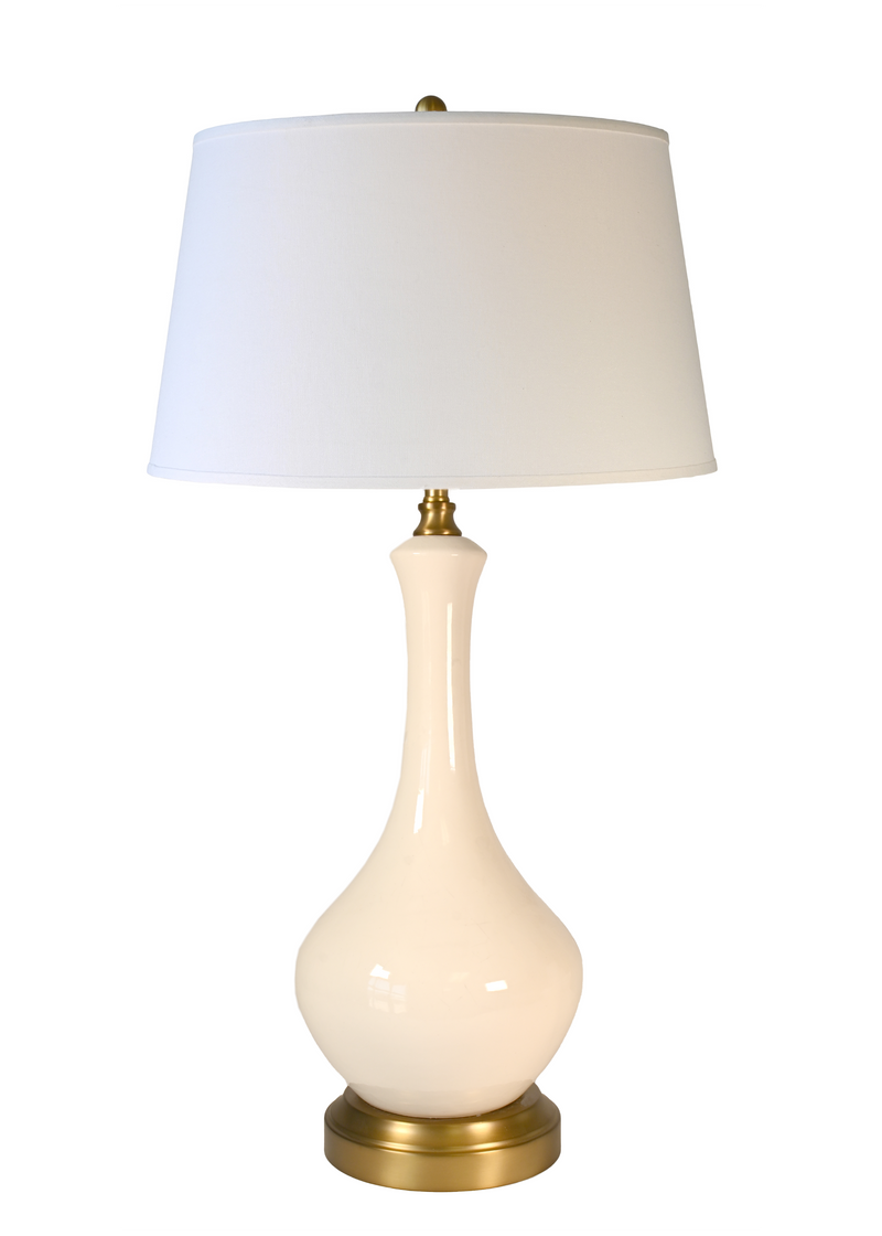 A cordless table lamp with a glossy cream-colored ceramic vase-shaped base, an off white fabric drum shade, and brass-finished metal accents at the top and bottom.