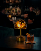 A warm, glowing cordless mini table lamp with a leopard-print glass dome and brass base sits on a dark bar table next to a glass of amber cocktail 